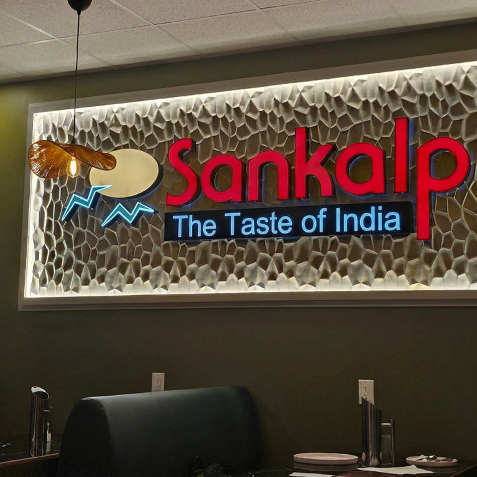 Illuminated Sankalp The Taste of India sign above dark restaurant booths, with decorative pendant lights and textured backdrop.